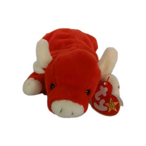 May include: A red plush toy bull with white horns, hooves, and muzzle. The stuffed animal has black eyes and a tag attached to its ear. The tag reads "Beanie Babies" and features a heart shape.