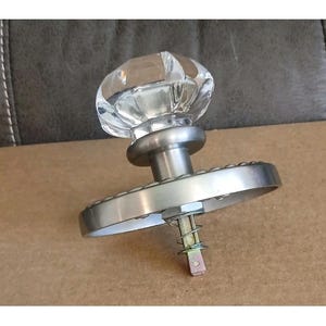 May include: A clear, faceted glass door knob with a brushed nickel base and backplate. The knob has a classic, vintage design. The backplate is round with a decorative edge. The hardware includes a spring and a square spindle.