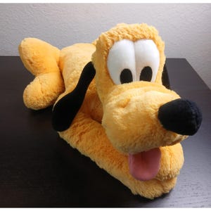 May include: A plush toy of Pluto, a yellow dog with black ears, a black nose, and a pink tongue. The dog's eyes are white with black pupils. The toy is lying down on a dark surface.