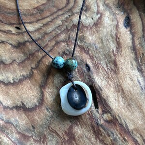 Könnte beinhalten: Eine Halskette mit einer braunen Schnur, zwei türkisfarbenen Perlen und einem weißen und schwarzen Anhänger. Der Anhänger ist ein rundes, flaches Stück Muschel mit einem kleineren, runden, schwarzen Stein im Inneren.