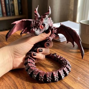 Può includere: Una statuetta di drago articolata rosso scuro e nera con un corpo squamoso e testurizzato, corna affilate e occhi rossi prominenti. La creatura presenta una lunga coda segmentata che si arriccia in forma circolare, con ali spiegate e punte dettagliate.