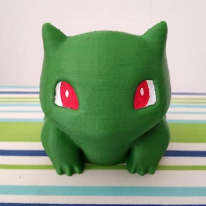May include: A green 3D printed figurine of a Pokemon character with red eyes. The figurine is sitting on a striped surface.