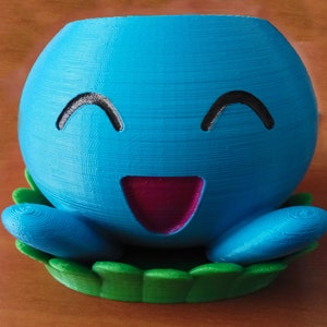May include: A blue 3D printed planter shaped like a cartoon character with a smiling face and a pink tongue. The planter is sitting on a green base that resembles grass.