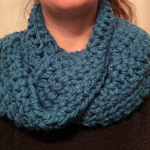 Könnte beinhalten: Ein teal-blauer, grob gestrickter Loop-Schal. Der Schal ist aus dickem Garn gefertigt, wodurch ein strukturiertes Aussehen entsteht. Der kreisförmige Schal wird um Hals und Schultern getragen. Dieses handgefertigte Accessoire ist perfekt für kaltes Wetter.