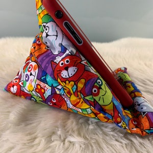 Könnte beinhalten: Ein rotes Smartphone liegt auf einem Stoff-Handy-Ständer. Der Ständer hat ein helles Muster aus Cartoon-Katzen in verschiedenen Farben. Der Hintergrund ist eine weiche, weiße, flauschige Oberfläche.