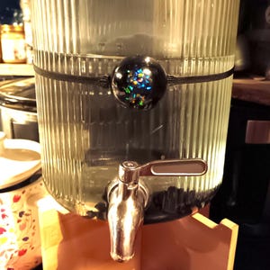 May include: A clear glass dispenser with a black rubber band and a black and iridescent round bottle stopper. The dispenser has a silver spigot with a lever handle.