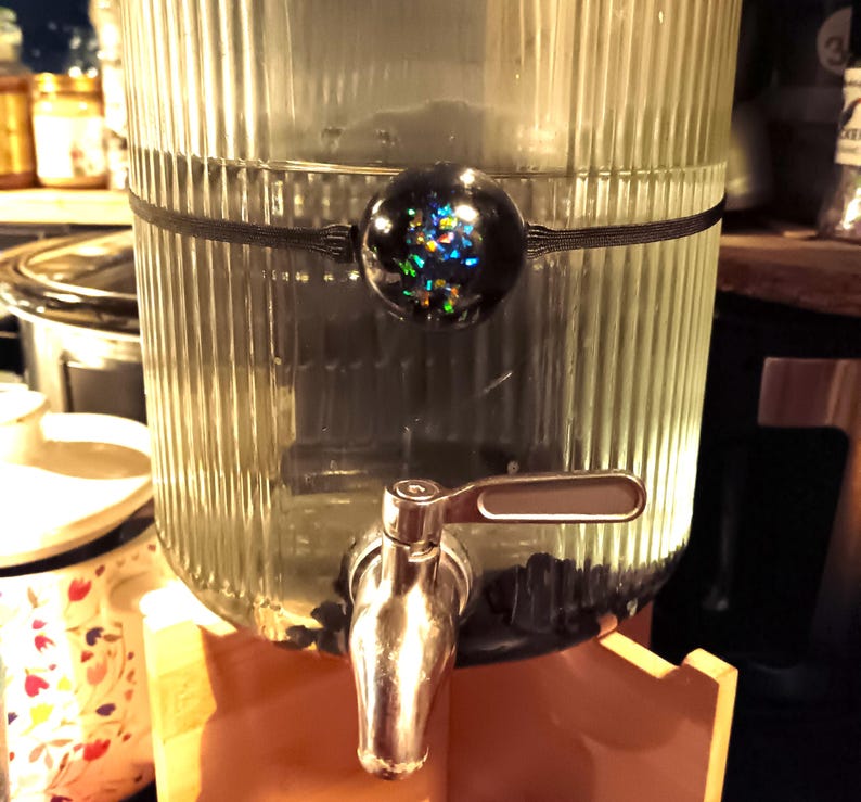 May include: A clear glass dispenser with a black rubber band and a black and iridescent round bottle stopper. The dispenser has a silver spigot with a lever handle.