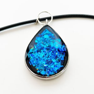 May include: A teardrop-shaped pendant necklace with a silver setting and a blue iridescent resin inlay. The necklace has a black cord.