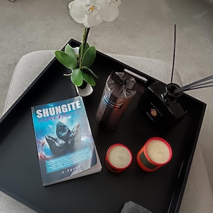 May include: A black tray with a book titled "Shungite: Inspect the Mystery", two red candles, a black bottle with a diffuser, a brown package, and a white orchid plant.