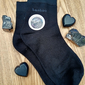 May include: A pair of black bamboo socks with the words "bamboo" and "Graphite Shungite" printed on them. The socks are lying on a wooden surface with black heart-shaped stones.