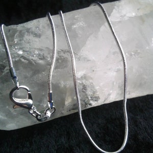 May include: A silver snake chain necklace with a lobster clasp closure. The chain is lying on a white crystal surface.