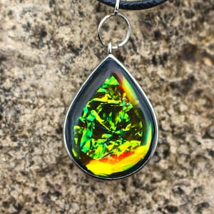 May include: A teardrop-shaped pendant necklace with a green, yellow, and orange iridescent glass insert. The pendant is attached to a black cord.