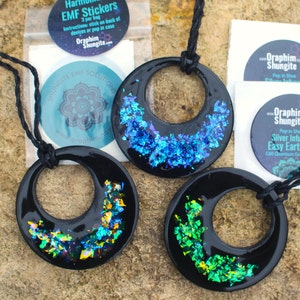 May include: Three black pendant necklaces with a circular design. Each pendant has a different color of iridescent glitter embedded in the resin. The text on the stickers reads "Oraphim Shungite EMF Solutions" and "Pop in Shoe Silver Infused Easy Earth C60 Quantum Grade".