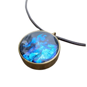 May include: A round pendant necklace with a dark brown cord. The pendant has a gold-colored bezel and a clear, glossy center filled with blue and black iridescent material. The pendant is approximately 1.5 inches in diameter.