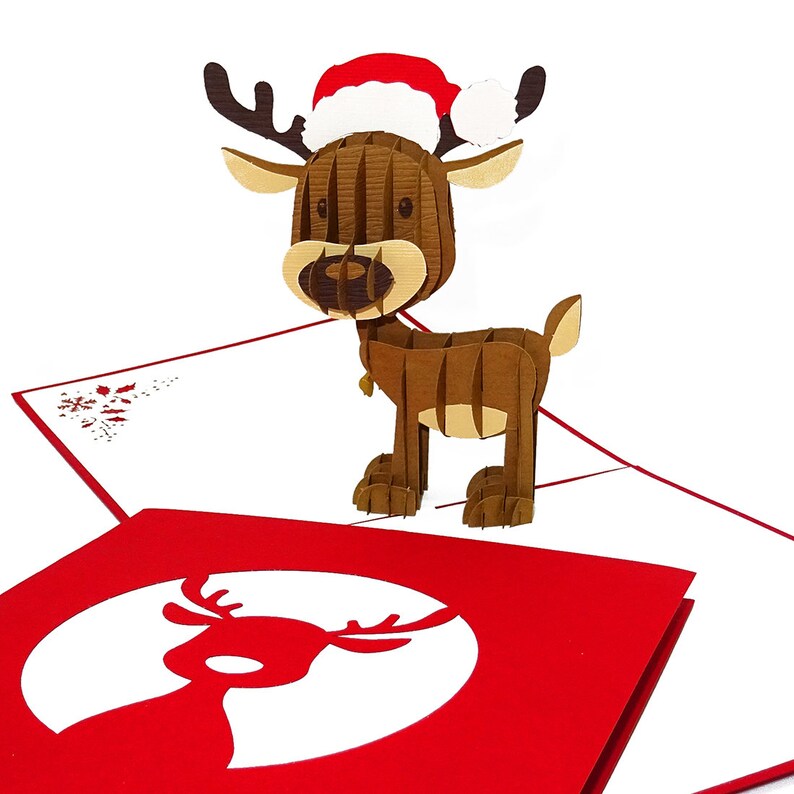 May include: A 3D pop-up Christmas card featuring a reindeer wearing a red Santa hat. The card is open and the reindeer is standing on a red background with a white reindeer silhouette.
