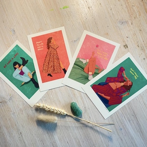 Könnte beinhalten: Vier Kunstdrucke mit Illustrationen und Text auf einer hellen Holzoberfläche. Die Drucke zeigen Frauen in verschiedenen Posen, mit Sätzen wie "Sit Like a Lady" und "Don't Forget You're Free." Ein Weizenähre und ein grüner Stein sind ebenfalls vorhanden.