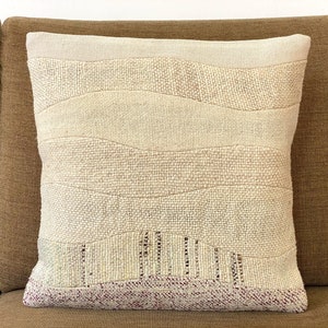 Peut inclure: Coussin décoratif crème avec un design texturé et superposé. Le coussin présente des bandes horizontales de différentes nuances de crème et de beige, avec un tissage texturé. Le bord inférieur a une bande bordeaux. Le coussin est sur un canapé marron.