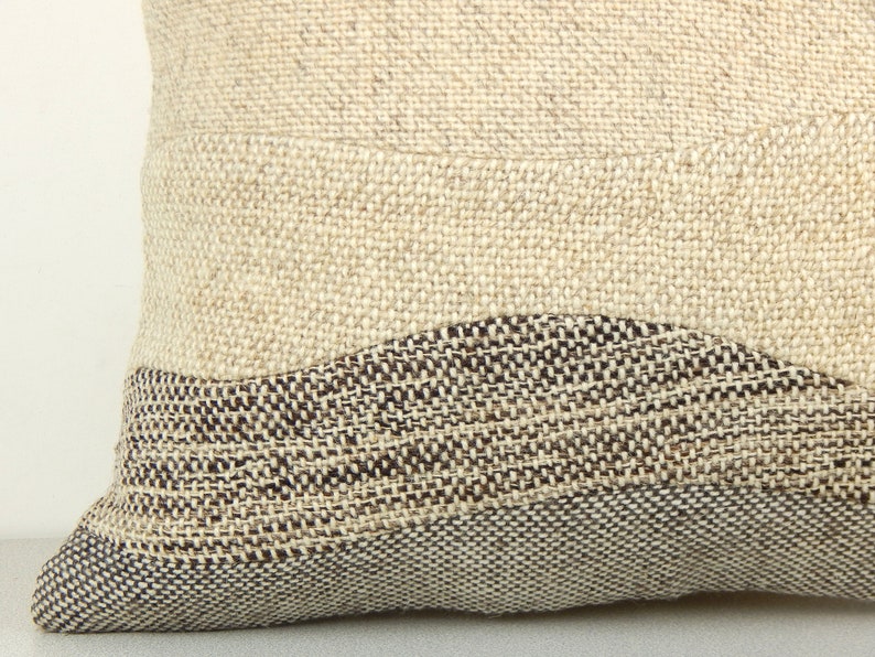 Op de afbeelding: Close-up van een getextureerd kussen met een neutraal kleurenpalet. Het kussen heeft een geweven ontwerp met een lichtbeige achtergrond en een donkerbruin, golvend patroon aan de onderkant. De stof lijkt een natuurlijke vezel te zijn.