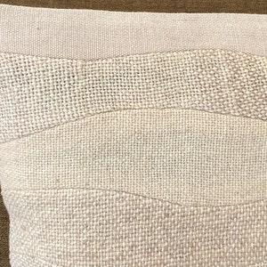 May include: Close-up of a beige decorative pillow with a textured, woven design. The pillow features horizontal bands of varying textures and shades of beige, creating a subtle, layered effect. The pillow's design is reminiscent of a landscape.