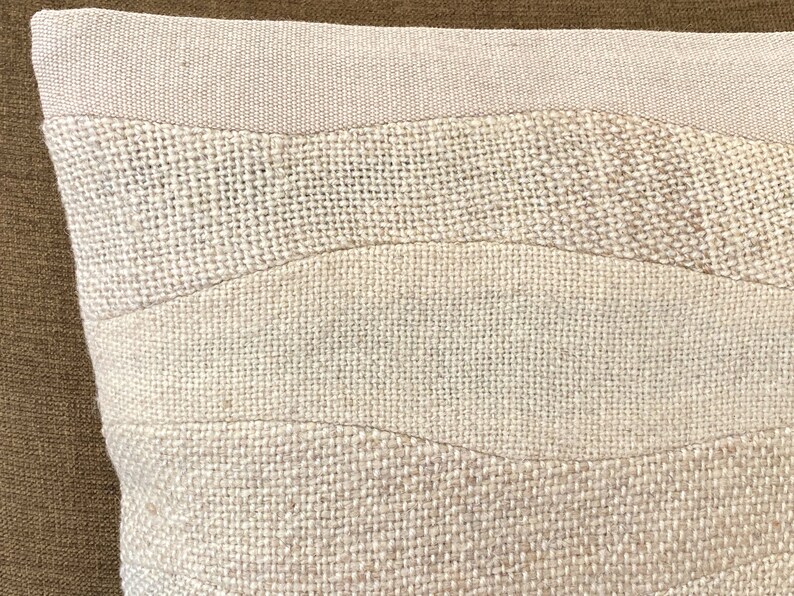 May include: Close-up of a beige decorative pillow with a textured, woven design. The pillow features horizontal bands of varying textures and shades of beige, creating a subtle, layered effect. The pillow's design is reminiscent of a landscape.