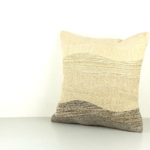 Op de afbeelding: Een vierkant decoratief kussen met een getextureerd, gelaagd ontwerp. Het kussen heeft een kleurengradi&euml;nt van lichtbeige tot donkergrijs, met horizontale lijnen die een golfachtig patroon cre&euml;ren. Het kussen is gemaakt van geweven stof.