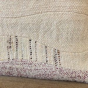 May include: Close-up of a beige decorative pillow with a textured, woven fabric. The pillow features a patchwork design with a wavy pattern and a burgundy border. The pillow is made of natural materials and has a rustic aesthetic.