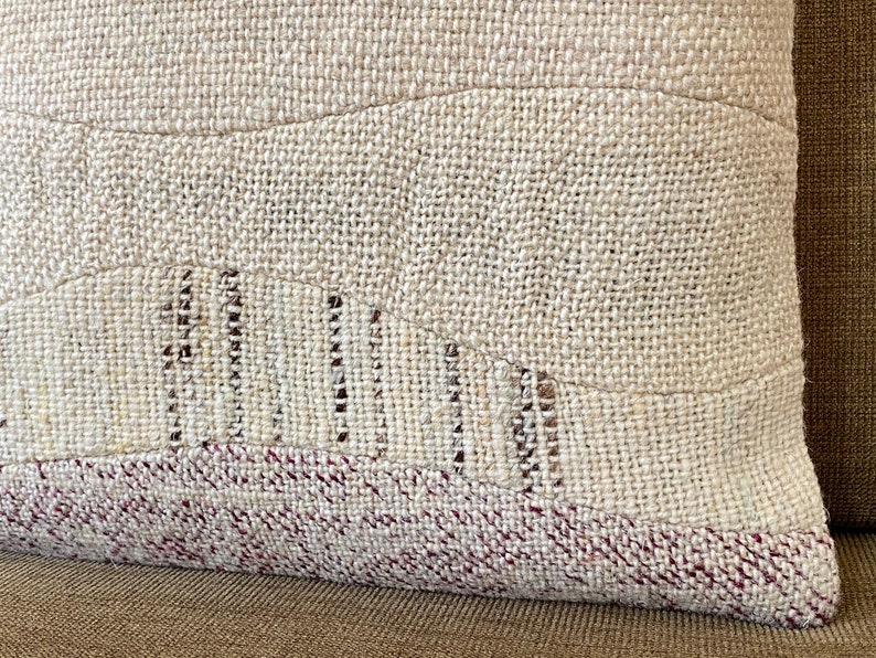 May include: Close-up of a beige decorative pillow with a textured, woven fabric. The pillow features a patchwork design with a wavy pattern and a burgundy border. The pillow is made of natural materials and has a rustic aesthetic.