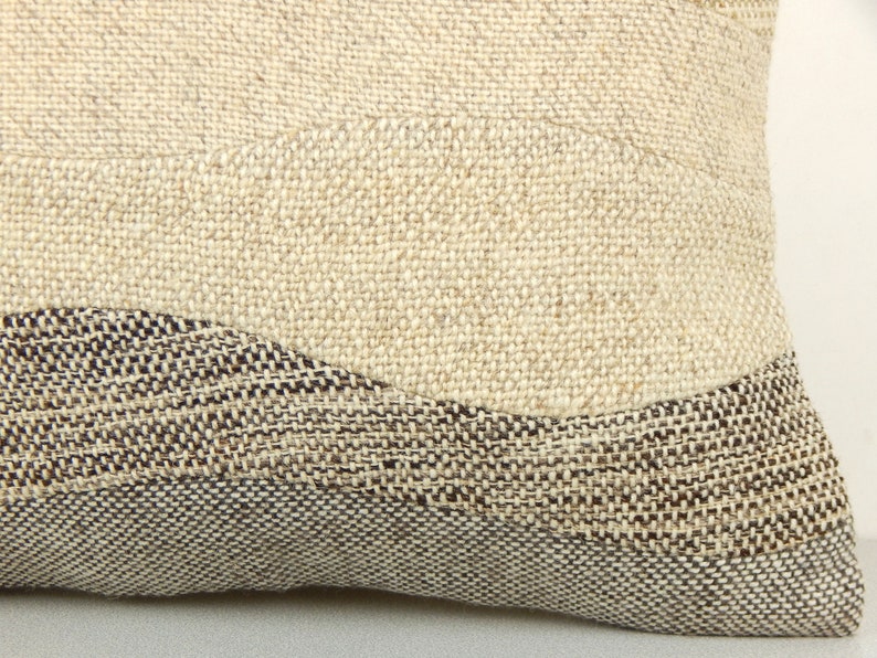 Op de afbeelding: Close-up van een beige en bruin geweven kussen. Het kussen heeft een textuur, neutraal getinte stof met een golfachtig ontwerp in donkerdere bruin- en beigetinten. Het ontwerp van het kussen is eenvoudig en modern, geschikt voor woondecoratie.
