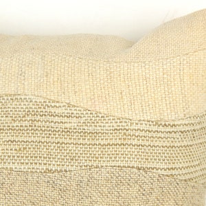 Op de afbeelding: Close-up van een beige decoratief kussen met een textuur, geweven ontwerp. Het kussen heeft horizontale banden van verschillende texturen en tinten beige, waardoor een gelaagd effect ontstaat. Het is een neutraal woondecoratie-item.