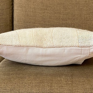 May include: A rectangular pillow with a patchwork design. The top section features a textured, off-white fabric with rectangular patches. The bottom section is a solid, light pink fabric. The pillow is resting on a brown surface.