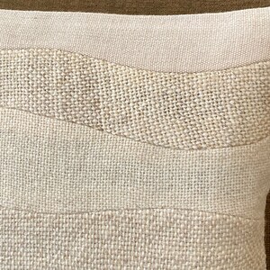 May include: Close-up of a beige decorative pillow with a textured, woven design. The pillow features horizontal bands of varying textures and shades of beige, creating a subtle, layered effect. The pillow's design adds a natural, earthy aesthetic.