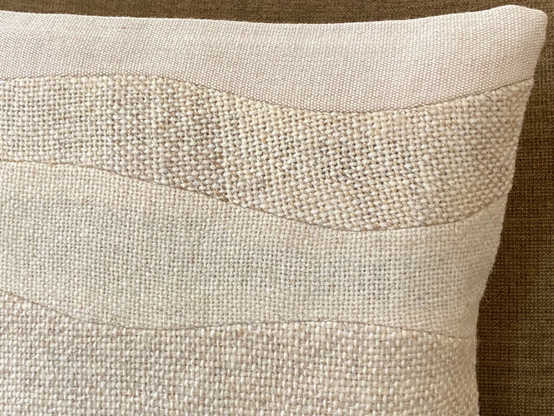 May include: Close-up of a beige decorative pillow with a textured, woven design. The pillow features horizontal bands of varying textures and shades of beige, creating a subtle, layered effect. The pillow's design adds a natural, earthy aesthetic.