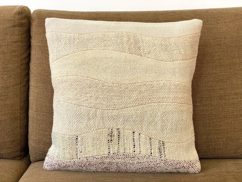 May include: Cream-colored decorative pillow with a textured, layered design. The pillow features horizontal bands of varying shades of cream and beige, with a textured weave. The bottom edge has a band of burgundy. The pillow is on a brown sofa.