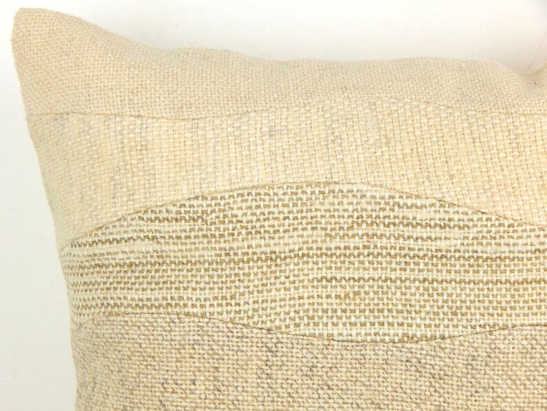 Op de afbeelding: Close-up van een beige decoratief kussen met een textuur, geweven ontwerp. Het kussen heeft horizontale banden met verschillende texturen en tinten beige, waardoor een subtiel, gelaagd effect ontstaat. De neutrale tinten van het kussen suggereren een natuurlijke, rustieke esthetiek.