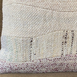 May include: Close-up of a textured pillow with a patchwork design. The pillow features a neutral color palette with beige and off-white tones. The bottom edge has a red and white woven pattern.