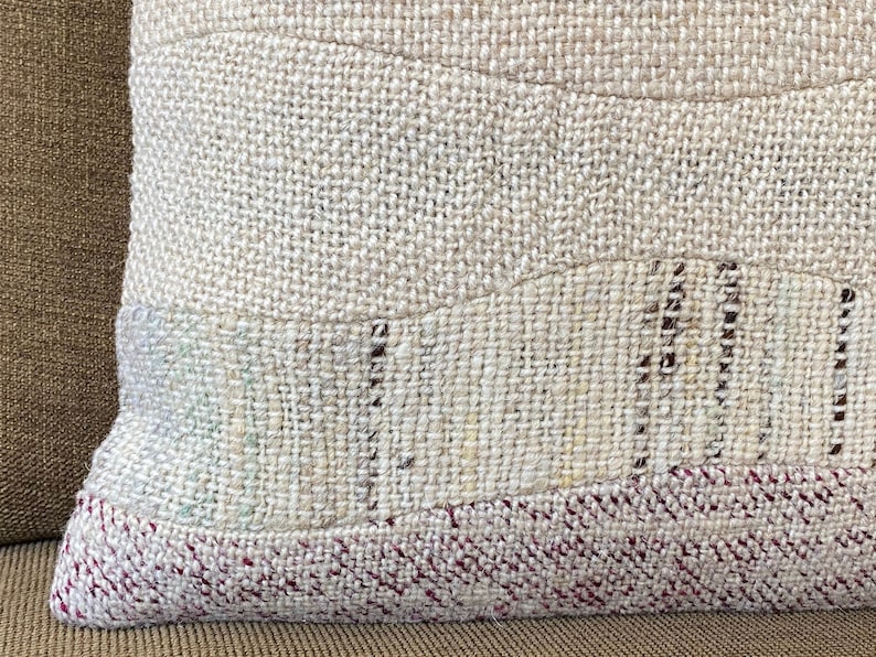 May include: Close-up of a textured pillow with a patchwork design. The pillow features a neutral color palette with beige and off-white tones. The bottom edge has a red and white woven pattern.