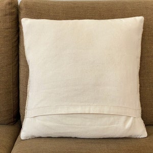 May include: A square, off-white pillow with a textured surface. The pillow is placed on a brown sofa. The pillow has a simple, minimalist design, suitable for home decor.