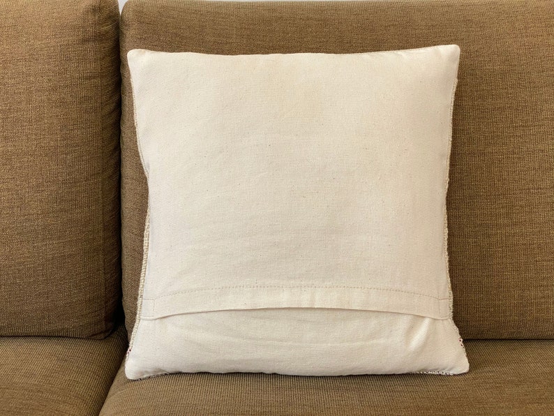May include: A square, off-white pillow with a textured surface. The pillow is placed on a brown sofa. The pillow has a simple, minimalist design, suitable for home decor.