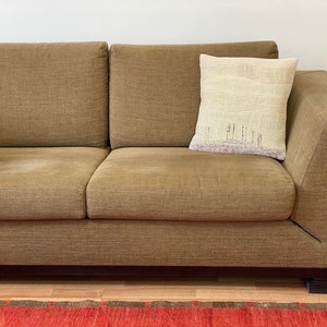 May include: A brown sectional sofa with a decorative pillow. The pillow is off-white with a landscape design in shades of beige and purple. The sofa has a modern design with a low profile and dark wooden feet.