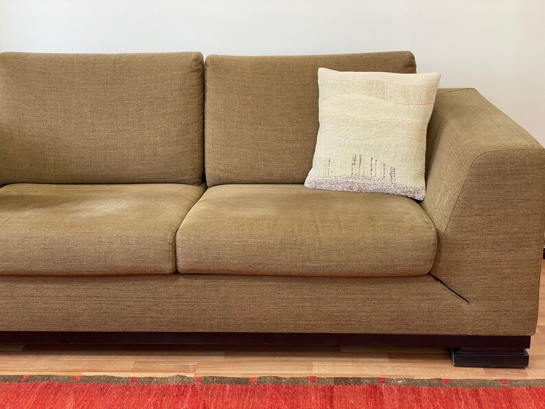 May include: A brown sectional sofa with a decorative pillow. The pillow is off-white with a landscape design in shades of beige and purple. The sofa has a modern design with a low profile and dark wooden feet.