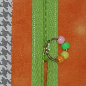 May include: A green zipper with a silver pull and a colorful beaded charm. The zipper is sewn into a patchwork fabric with a variety of patterns and colors.