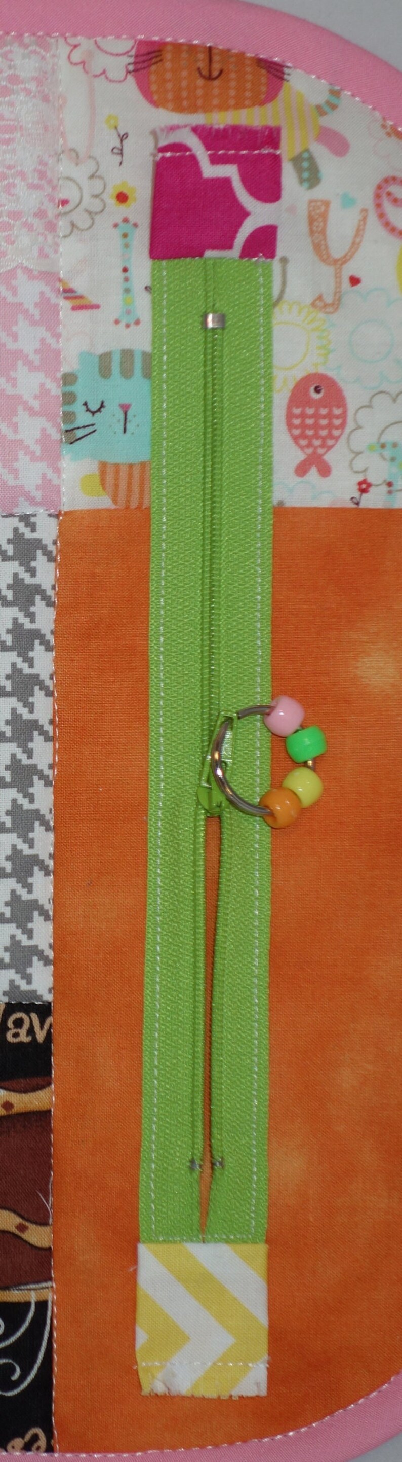 May include: A green zipper with a silver pull and a colorful beaded charm. The zipper is sewn into a patchwork fabric with a variety of patterns and colors.