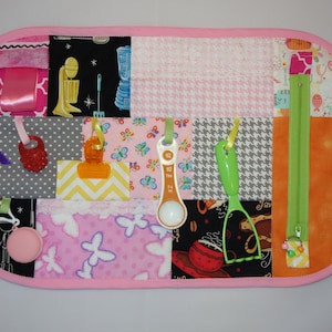 May include: A pink and gray fabric activity mat with various textures and objects for babies and toddlers to explore. The mat features a zipper, a measuring spoon, a whisk, a ribbon, a teether, and a clip.