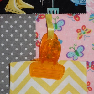 May include: A close-up of a yellow and white chevron fabric square attached to a pink fabric square with a bright orange plastic clip. The clip is attached to a yellow ribbon. The pink fabric square has a pattern of colorful butterflies.