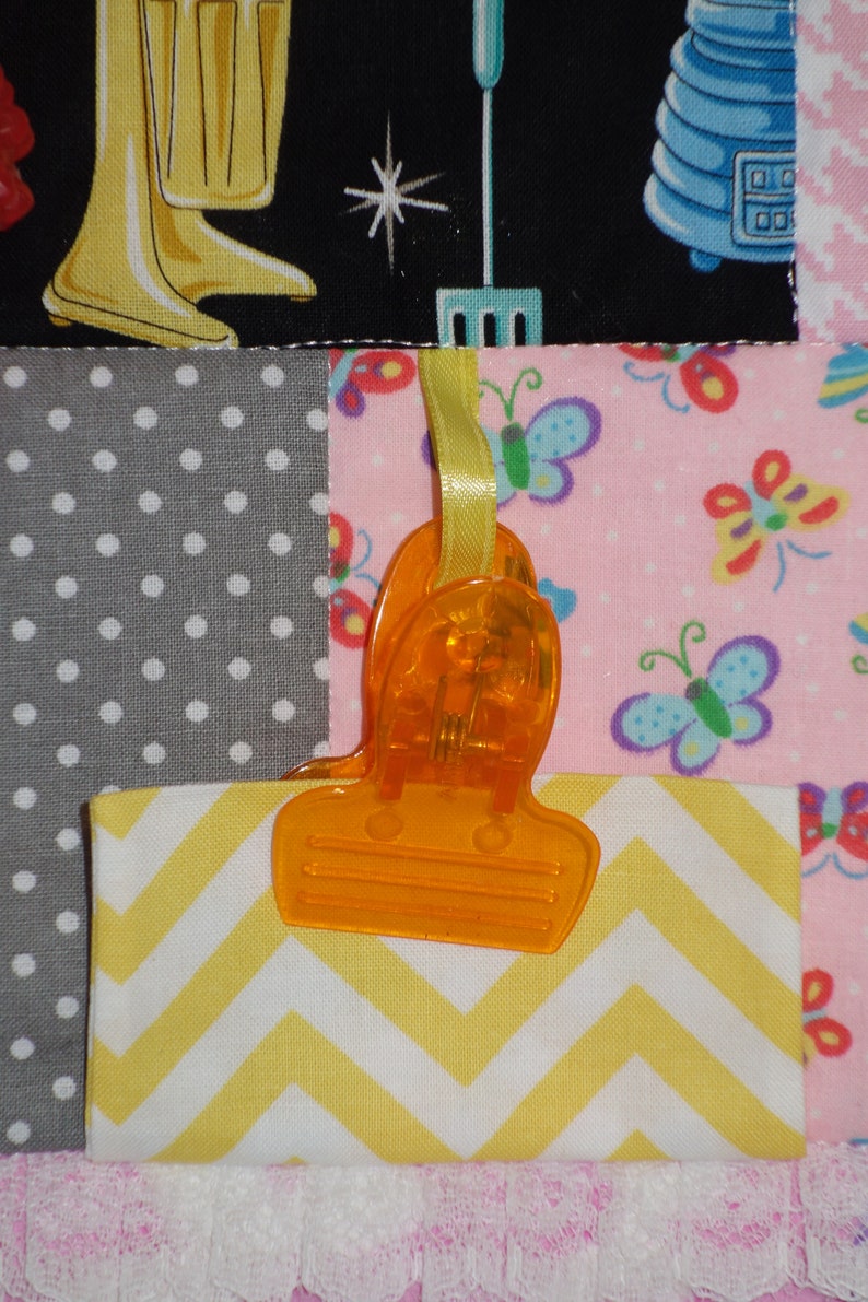 May include: A close-up of a yellow and white chevron fabric square attached to a pink fabric square with a bright orange plastic clip. The clip is attached to a yellow ribbon. The pink fabric square has a pattern of colorful butterflies.
