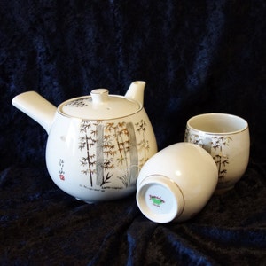 May include: A white ceramic teapot and two teacups with a gold bamboo design. The teapot has a spout and a handle. The teacups have a gold rim. The teapot and teacups are on a black background.
