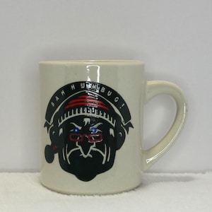 May include: White ceramic mug with a black and white cartoon character wearing a red and white hat and glasses. The text "Bah Humbug!" is written in a black circle around the character.
