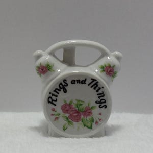May include: White ceramic alarm clock with a floral design and the words "Rings and Things" on the face.