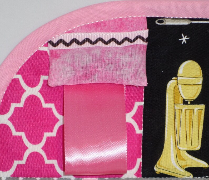 May include: Pink and white fabric with a geometric pattern. A pink ribbon is attached to the fabric. A pink and white fabric with a black zig-zag trim is attached to the fabric. A black and white fabric with a yellow mixer is attached to the fabric.