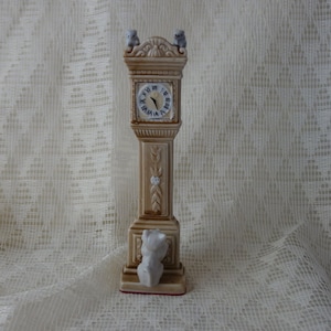 May include: A ceramic grandfather clock with a white face and black Roman numeral hour markers. Two small birds are perched on top of the clock. A white ceramic cat is sitting at the base of the clock.